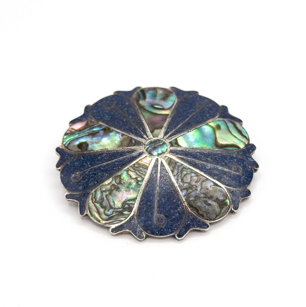 Face tilted towards the viewer on white, the small round abalone centre and the thin silver ribs radiating out between deep blue crushed stone wedges and iridescent shell segments.