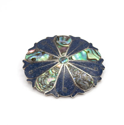 Face tilted towards the viewer on white, the small round abalone centre and the thin silver ribs radiating out between deep blue crushed stone wedges and iridescent shell segments.