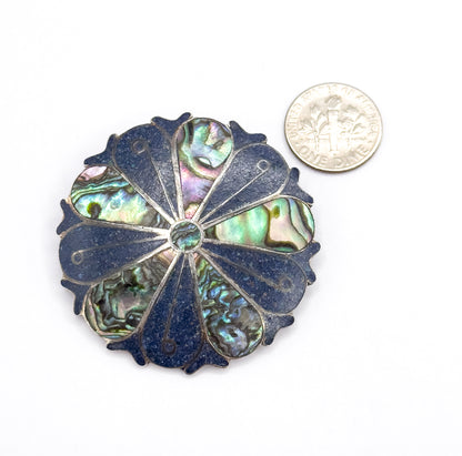 Round sterling silver pinwheel brooch with wedge sections of iridescent abalone alternating with dark crushed stone inlay, shown face-up on white beside a dime for scale.