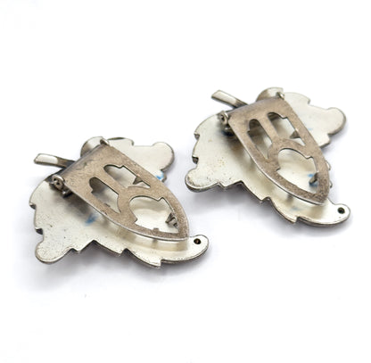 Angled from the side, the backs show the pierced shield-shaped plates standing proud of the leaf outlines with the hinged clip blades lifted away from the metal.