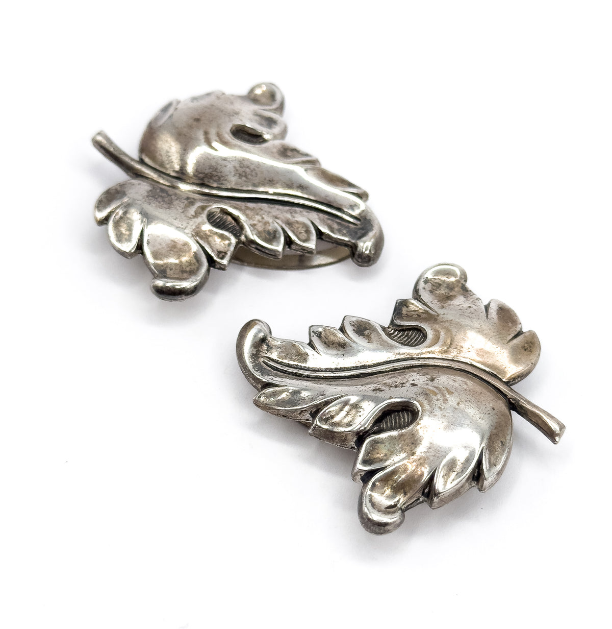 Both clips lie face-up on plain white, set diagonally so the curled scalloped edges, raised central stems and the narrow slot through each leaf front stand out against the soft patina.