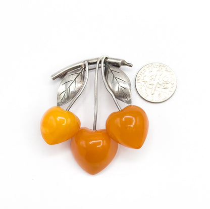 Laid flat face-up beside a small coin, which sits just under half the height of the brooch, giving scale to the three apricot amber cherries and the two veined silver leaves above them.