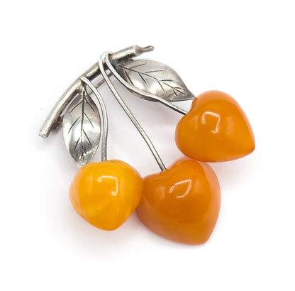 Overhead view on white, the three apricot amber cherries clustered at lower right while the silver stems curve up to two veined leaves with engraved midribs at upper left.