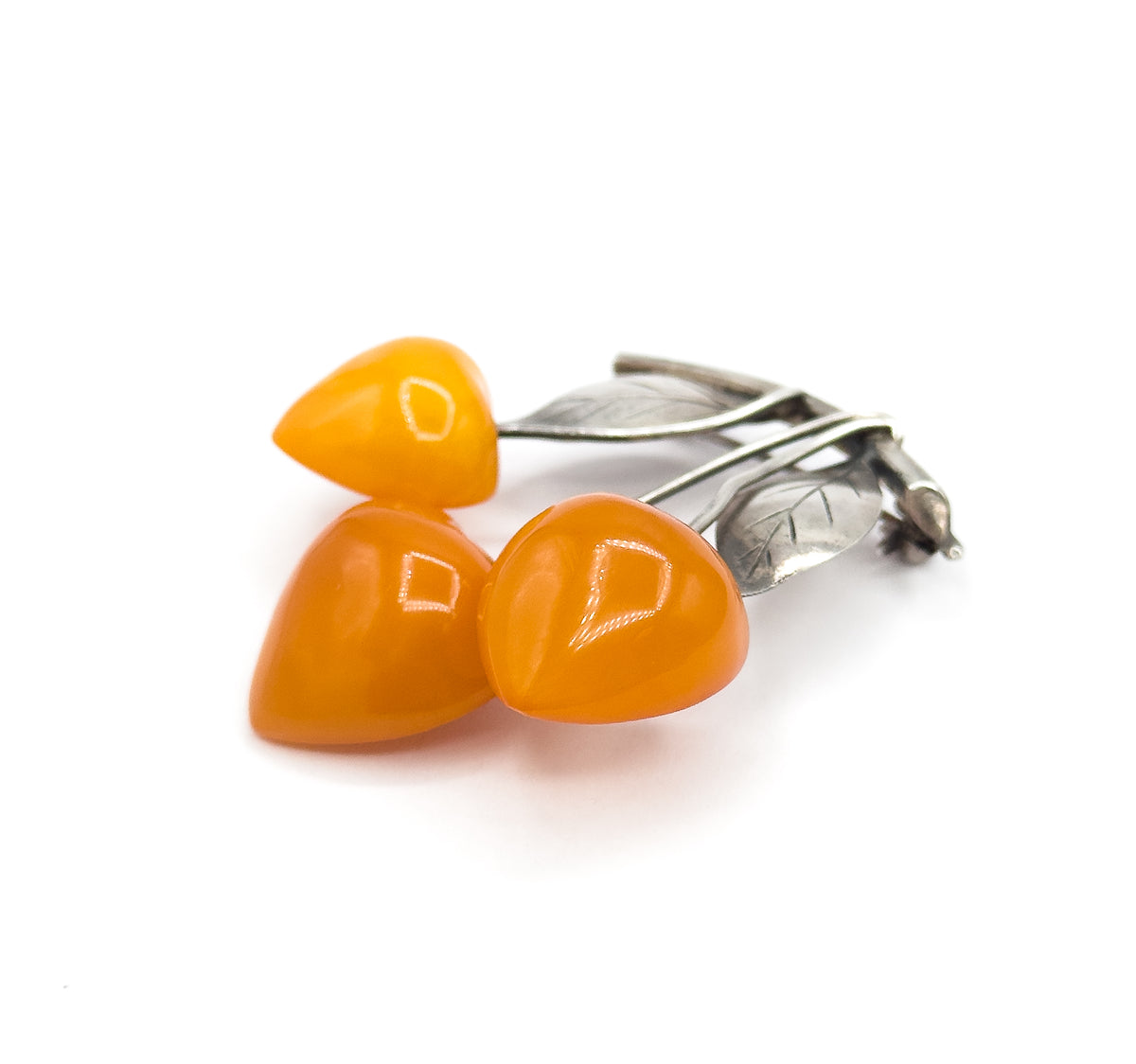 Low side view with the three domed apricot amber cherries clustered at the front, light glinting on their polished tops, while the silver leaves and the hook-shaped catch trail away to the right.