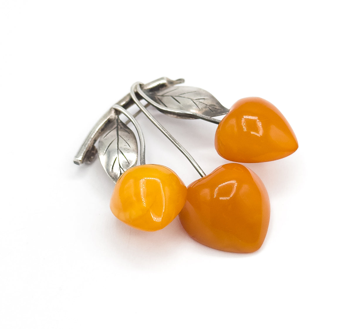 Close low three-quarter view with the three apricot amber cherries filling the foreground and their silver stems receding behind to the two veined leaves and the pin stem at the upper left.