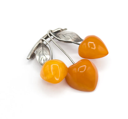 Close low three-quarter view with the three apricot amber cherries filling the foreground and their silver stems receding behind to the two veined leaves and the pin stem at the upper left.