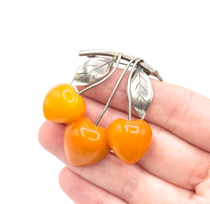 Resting across an open palm, the cherry brooch spans roughly three fingers, its three apricot amber fruits and two veined silver leaves giving a clear sense of scale against the hand.