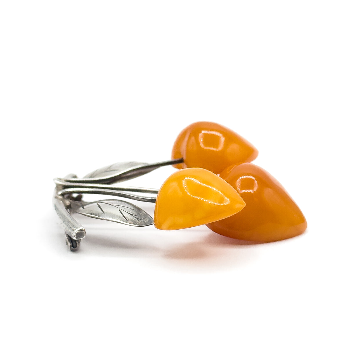 Cherry fruit amber brooch in 800 silver, shown at a low side angle on white, three polished apricot amber drops on silver stems with leaves, the pin stem and catch visible behind.