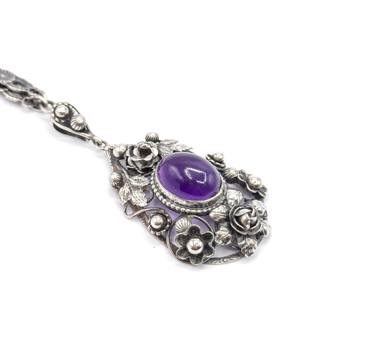 Angled three-quarter view down onto the pendant face, the amethyst in its beaded bezel ringed by roses, leaves and berries above the openwork lower gallery, bail and chain trailing up to the left.