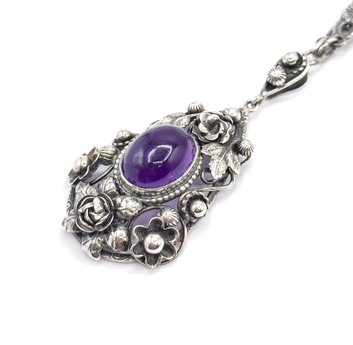 Diagonal view with the pendant at lower left and the chain running off to the upper right, showing the tapered bail with its applied blossom and the link joining it to the amethyst's beaded bezel.