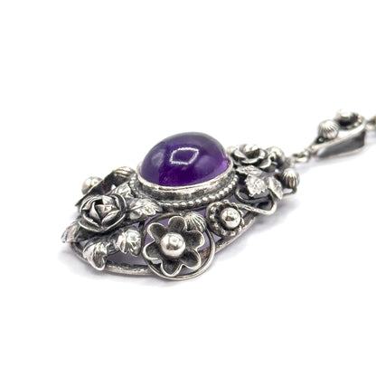 Tilted view from the lower edge, bringing the gallery's five-petalled flower and silver bead forward, the amethyst dome above it and the bail with its blossom angled away to the upper right.