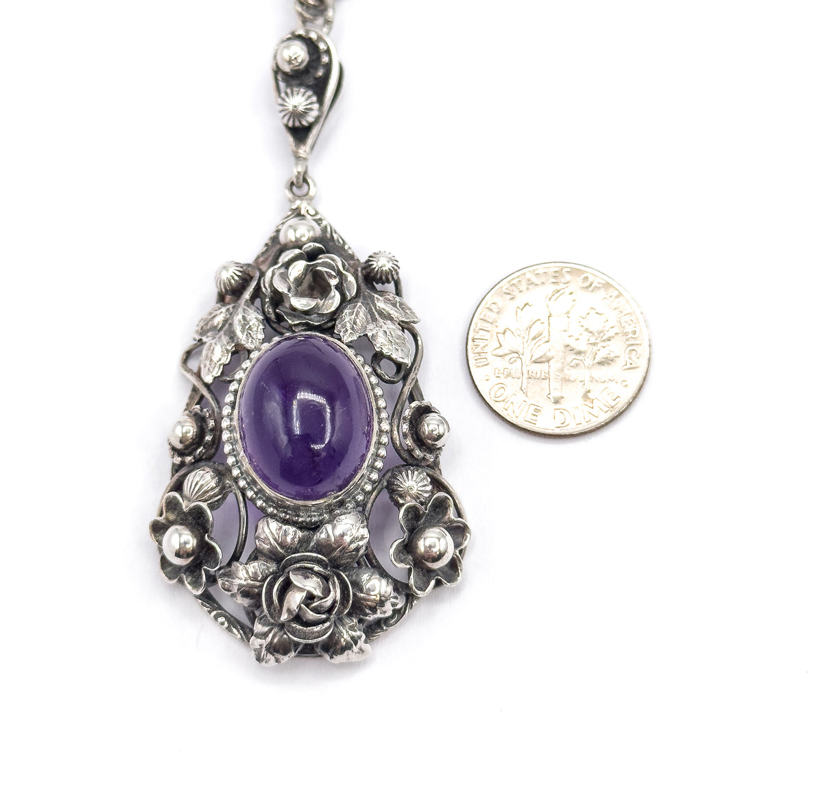 Flat front view beside a dime for scale, the coin roughly as wide as the amethyst's beaded bezel, with the whole floral sterling silver frame and bail visible above.