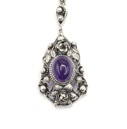 Straight-on upright view of the whole pendant hanging from its chain, the oval amethyst in a beaded bezel between a rose above and a rose below, with four silver blossoms at the sides.