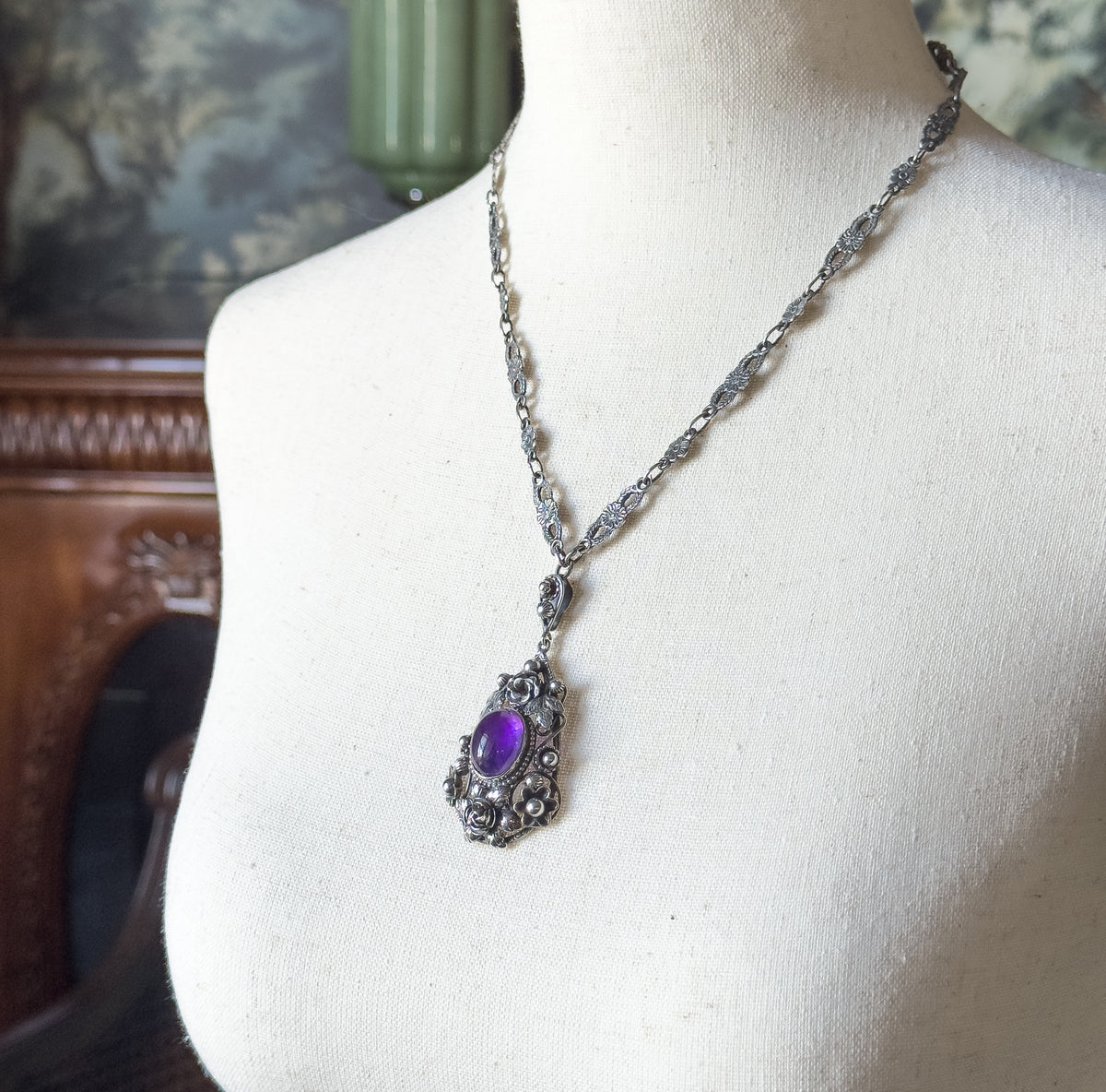 Arts and Crafts amethyst pendant in sterling silver, shown at the neck on a cream display form, the purple stone in its floral silver frame hanging from a patterned chain.