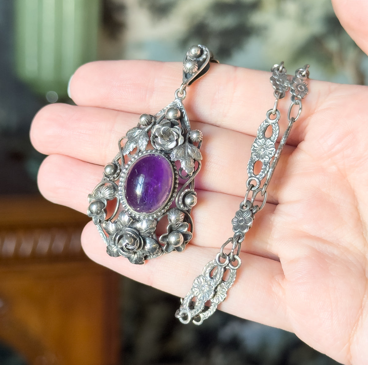 Shown resting on an open palm with the chain coiled beside it, giving a sense of scale: the oval purple amethyst in its beaded bezel surrounded by silver roses, buds and leaves.