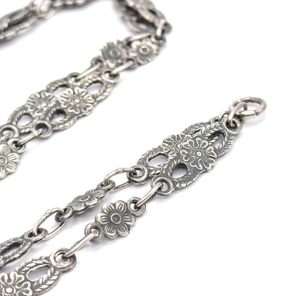 Close view of the sterling silver chain alone, its pierced oval panels of flowers and leaves alternating with small round blossom links and plain connecting rings, ending at a jump ring.