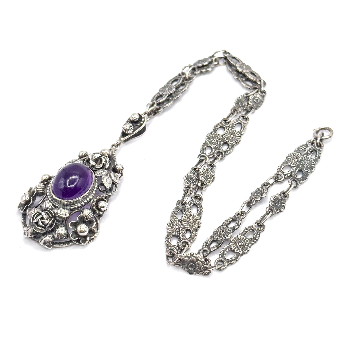 Laid flat on white with the chain looped to the right, the whole necklace seen from above: pendant at lower left, its amethyst bezel below a small bail joining the floral links.