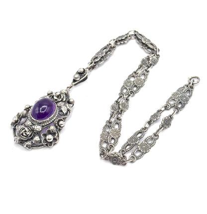 Laid flat on white with the chain looped to the right, the whole necklace seen from above: pendant at lower left, its amethyst bezel below a small bail joining the floral links.