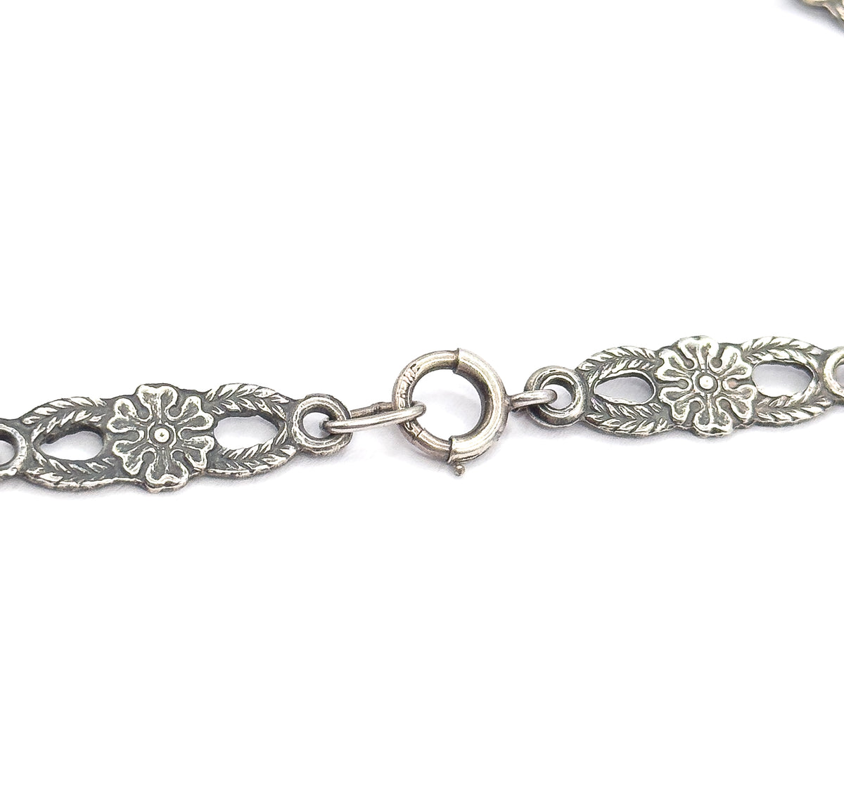 Close view of the round spring-ring clasp with its sliding tab, linked by small jump rings between two pierced floral chain panels, patina darkening the recesses.