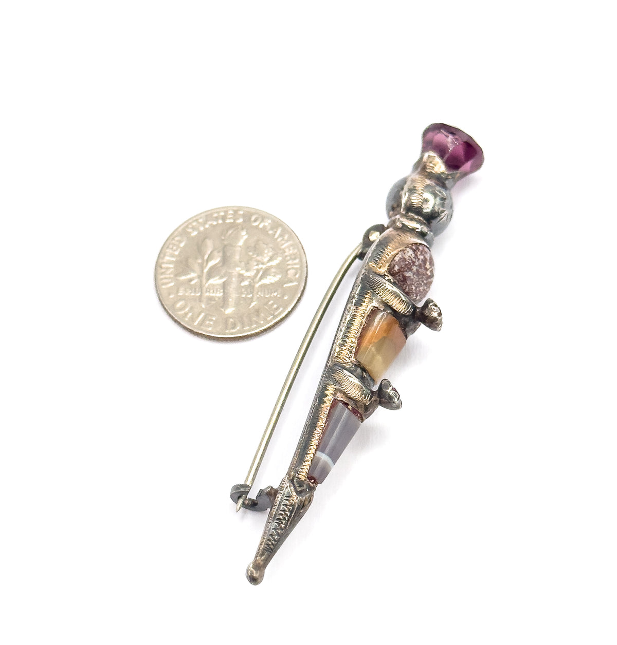 The brooch lies beside a dime for scale, back view showing the long pin stem and catch alongside the engraved silver body, purple paste at the thistle head.