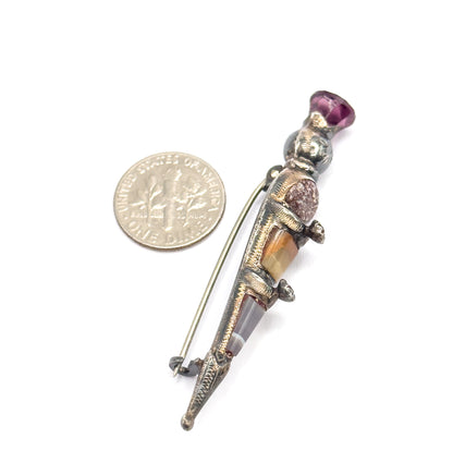 The brooch lies beside a dime for scale, back view showing the long pin stem and catch alongside the engraved silver body, purple paste at the thistle head.