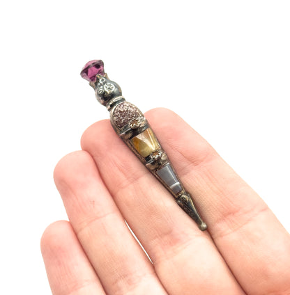 The brooch rests diagonally across open fingers, shown on the hand for scale, its length spanning barely more than two finger widths from pointed tip to purple paste thistle head.