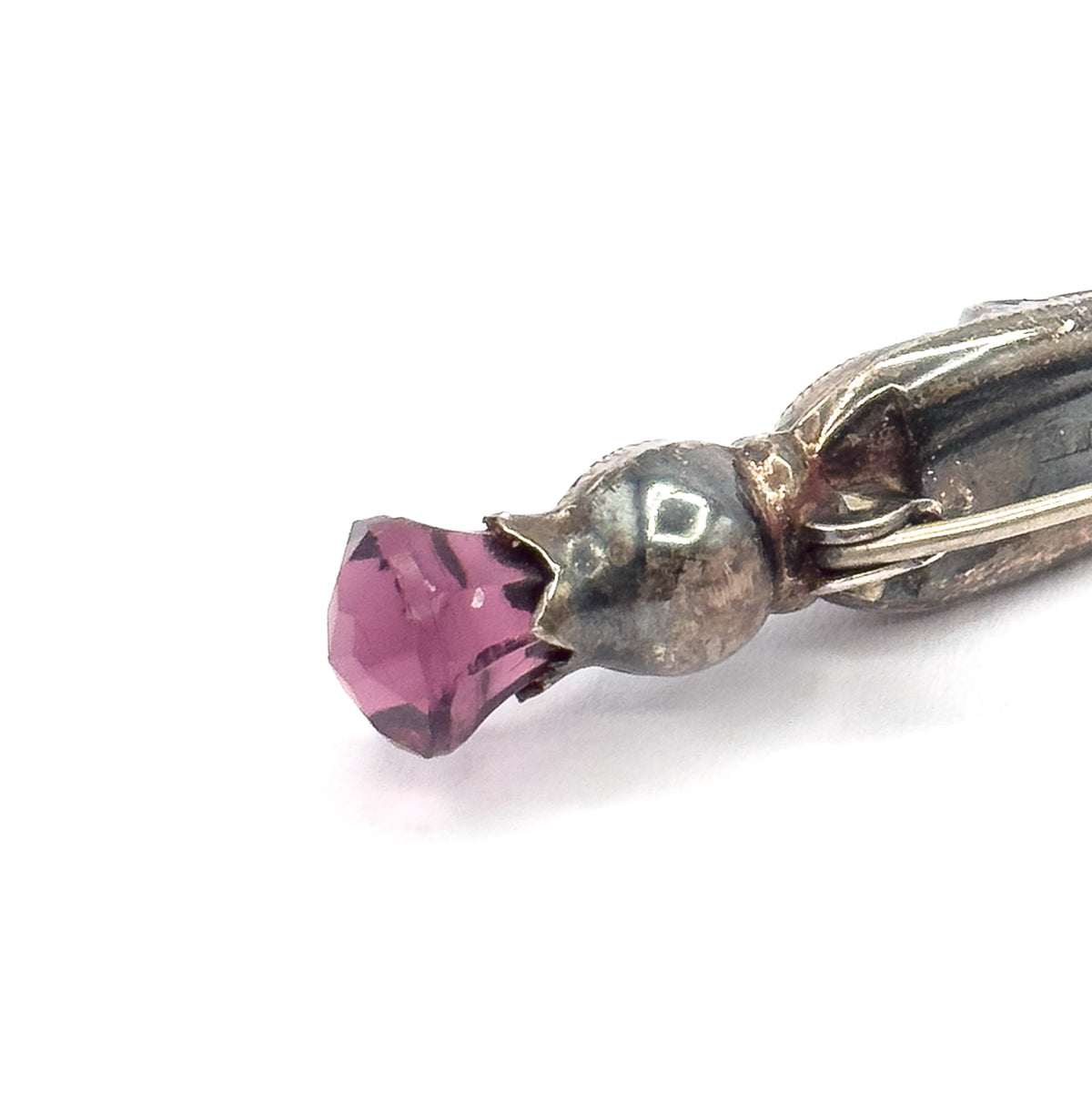Extreme close-up of the thistle head seen end-on, the faceted purple paste stone jutting from its pointed engraved cap, with the pin stem wire crossing the darkened patinated silver behind.