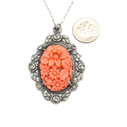 Shown beside a dime for scale, the pendant hanging from its chain with the coin roughly two-thirds the width of the oval frame of silver roses, leaves and marcasites.