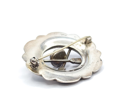 The reverse, showing the plain scalloped back with its pin stem crossing the open centre from hinge to catch, the curling stone motif visible through the opening and lettering stamped near the rim.