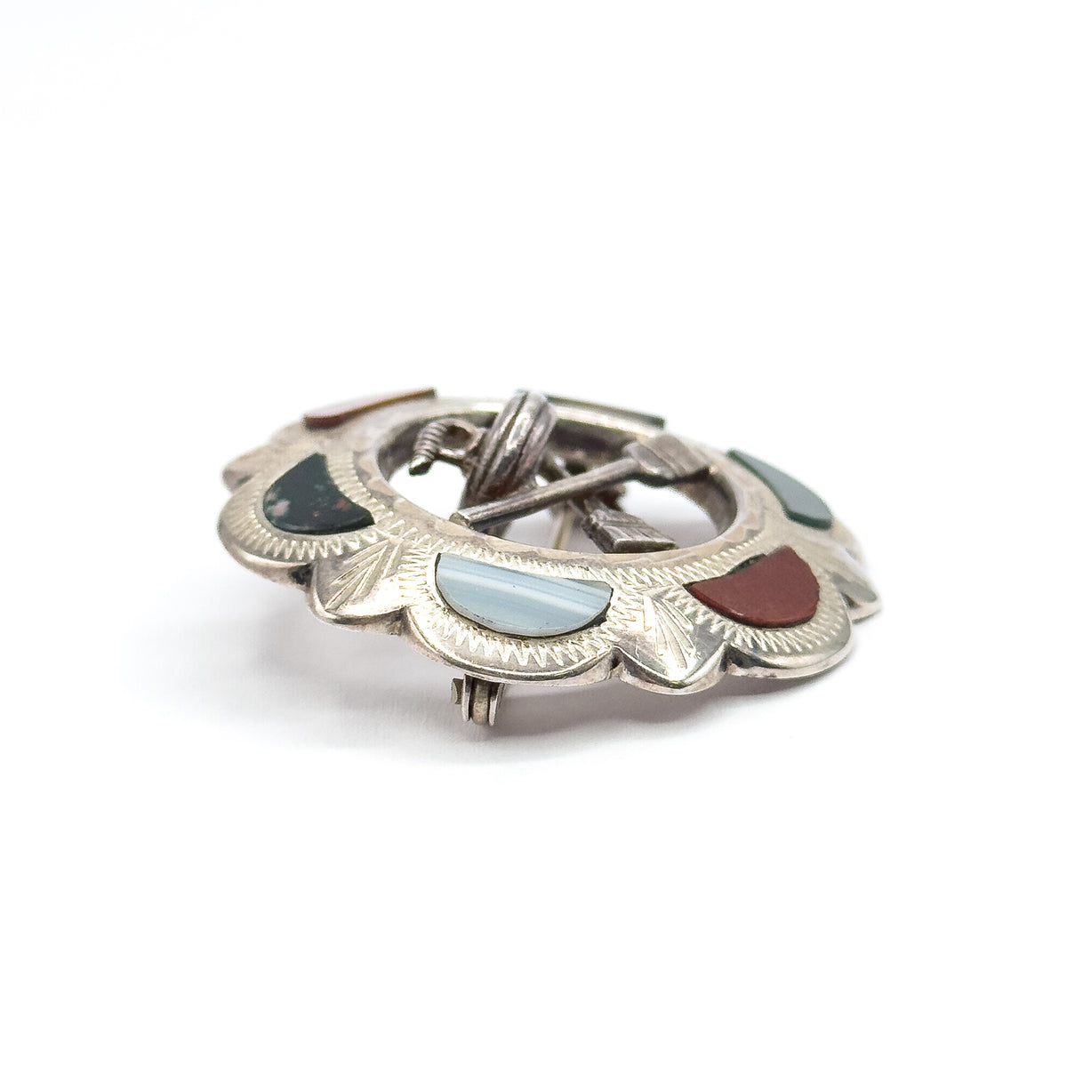 A near-level close view across the scalloped rim, where a banded pale blue cabochon, a dark green one and a red one sit in engraved panels, with the hinge and pin stem rising at the centre.