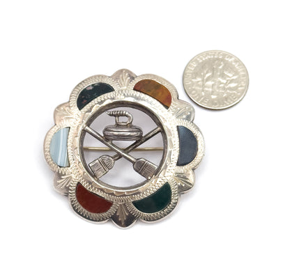 The front shown flat beside a dime for scale, the curling stone and crossed brooms filling the open centre within a scalloped border of green, orange, blue, grey and red cabochons.