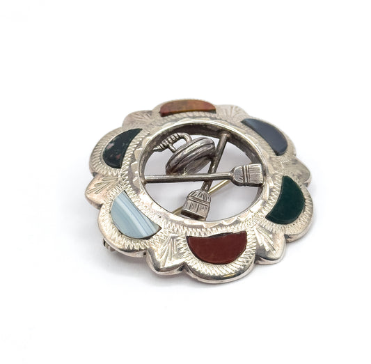 A tilted overhead view showing the whole ring of cabochons around the scalloped border at once, with the pin stem crossing diagonally through the open centre past the curling stone and brooms.