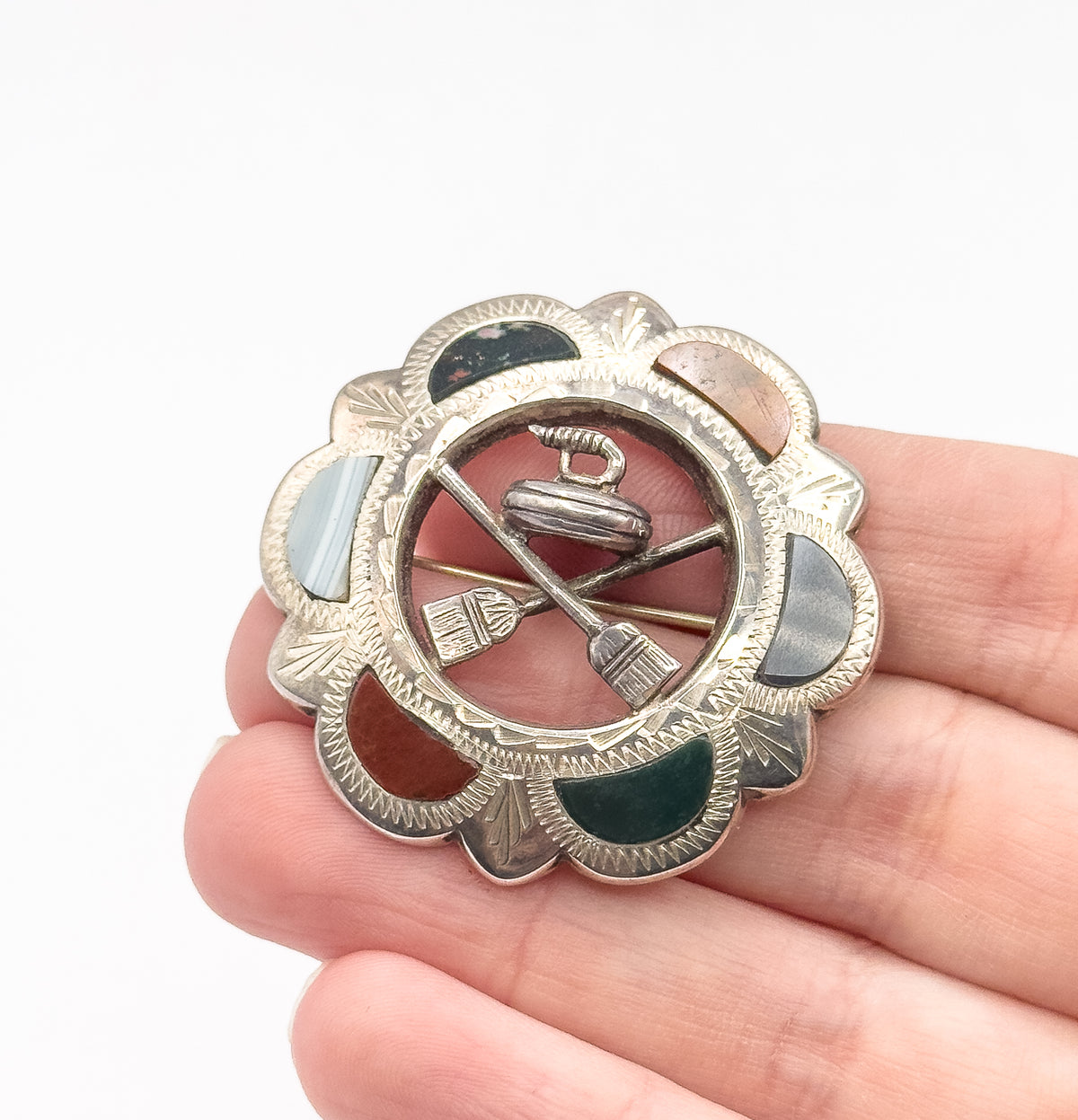 Vintage Scottish Celtic shield brooch in sterling silver, shown resting on open fingertips, its scalloped border set with coloured glass cabochons around a curling stone and crossed brooms.