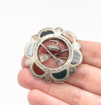 Vintage Scottish Celtic shield brooch in sterling silver, shown resting on open fingertips, its scalloped border set with coloured glass cabochons around a curling stone and crossed brooms.