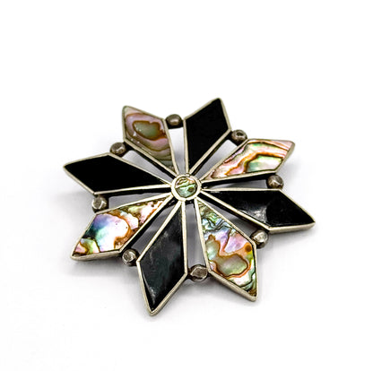 The face tilted at an angle on white, showing eight diamond-shaped points of black onyx and iridescent abalone radiating from a small round abalone centre, silver balls between each.