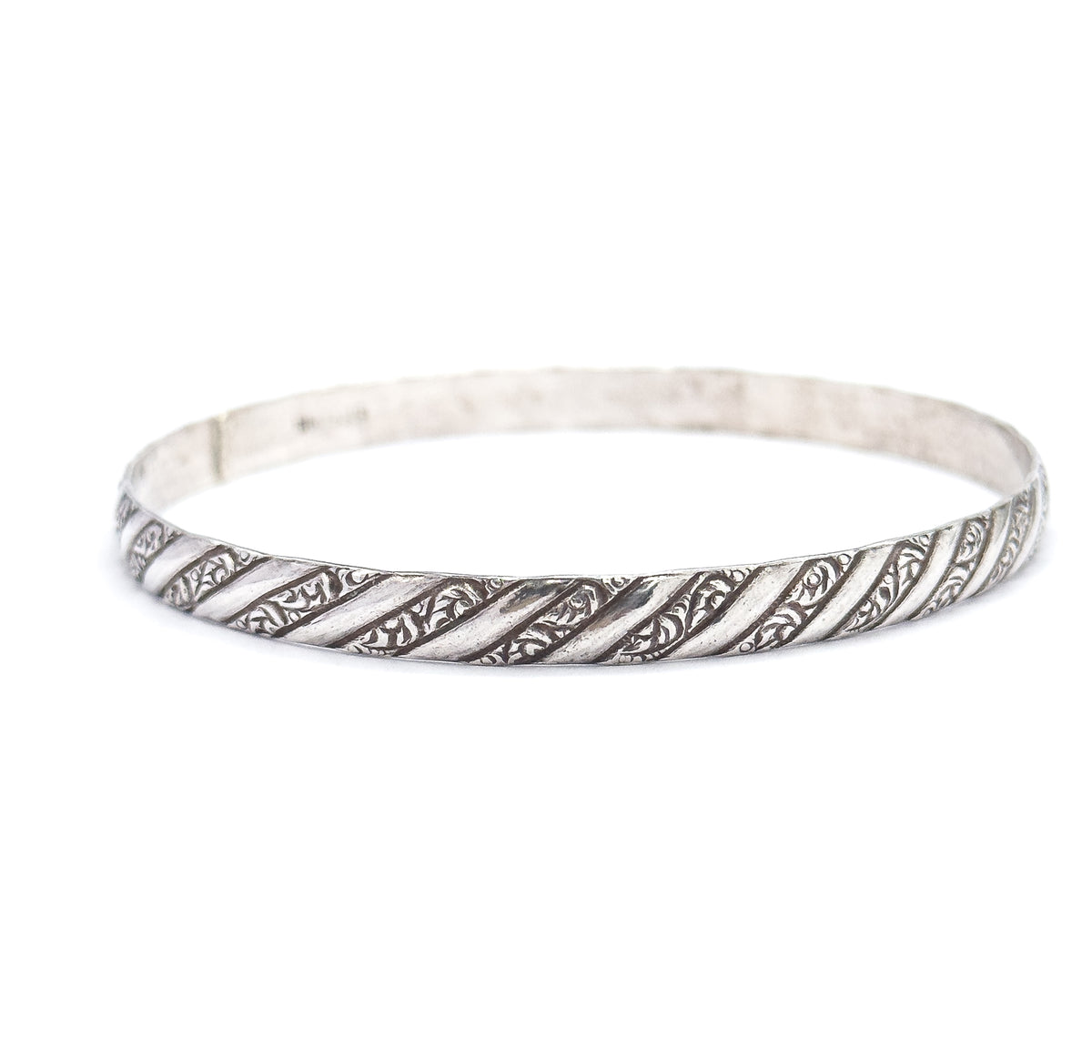 The bangle lies tilted low and nearly edge-on, its smooth inner wall and faint stamped mark visible at the back while the repousse stripes run unbroken along the front rim.