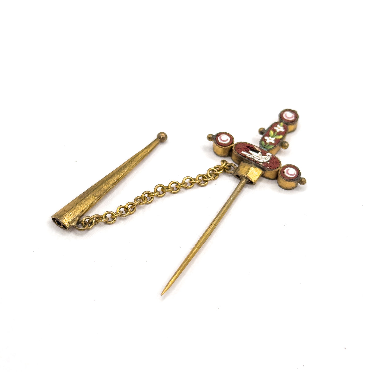 The sword pin lies flat on white, long pin stem pointing down from the mosaic hilt, with the tapered sheath set apart to the left and joined by its chain.