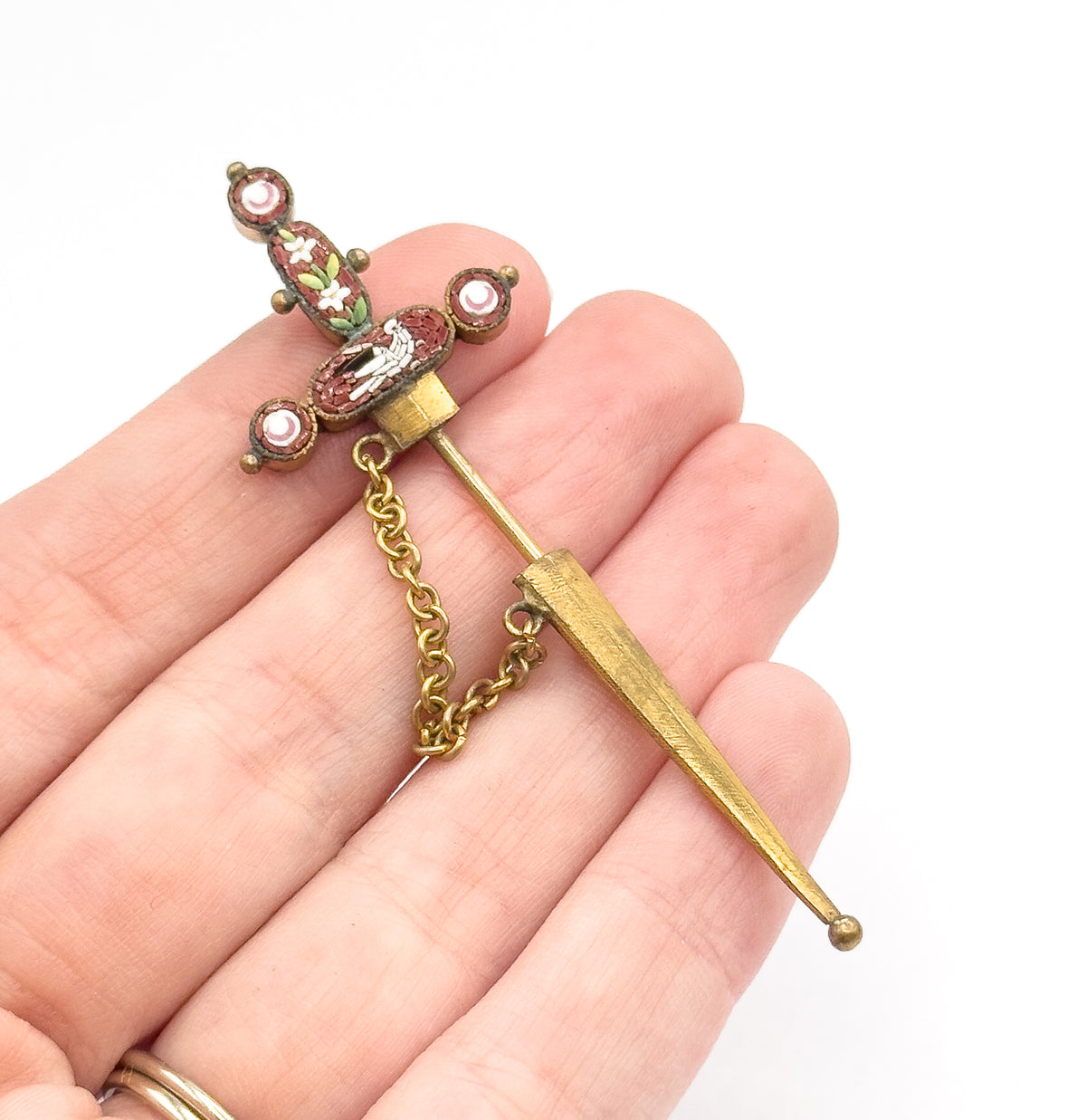 Base metal sword stick pin with rust-toned micro mosaic hilt, white dove and flowers, shown resting across open fingers with its sheath and small linking chain, giving a sense of its size.
