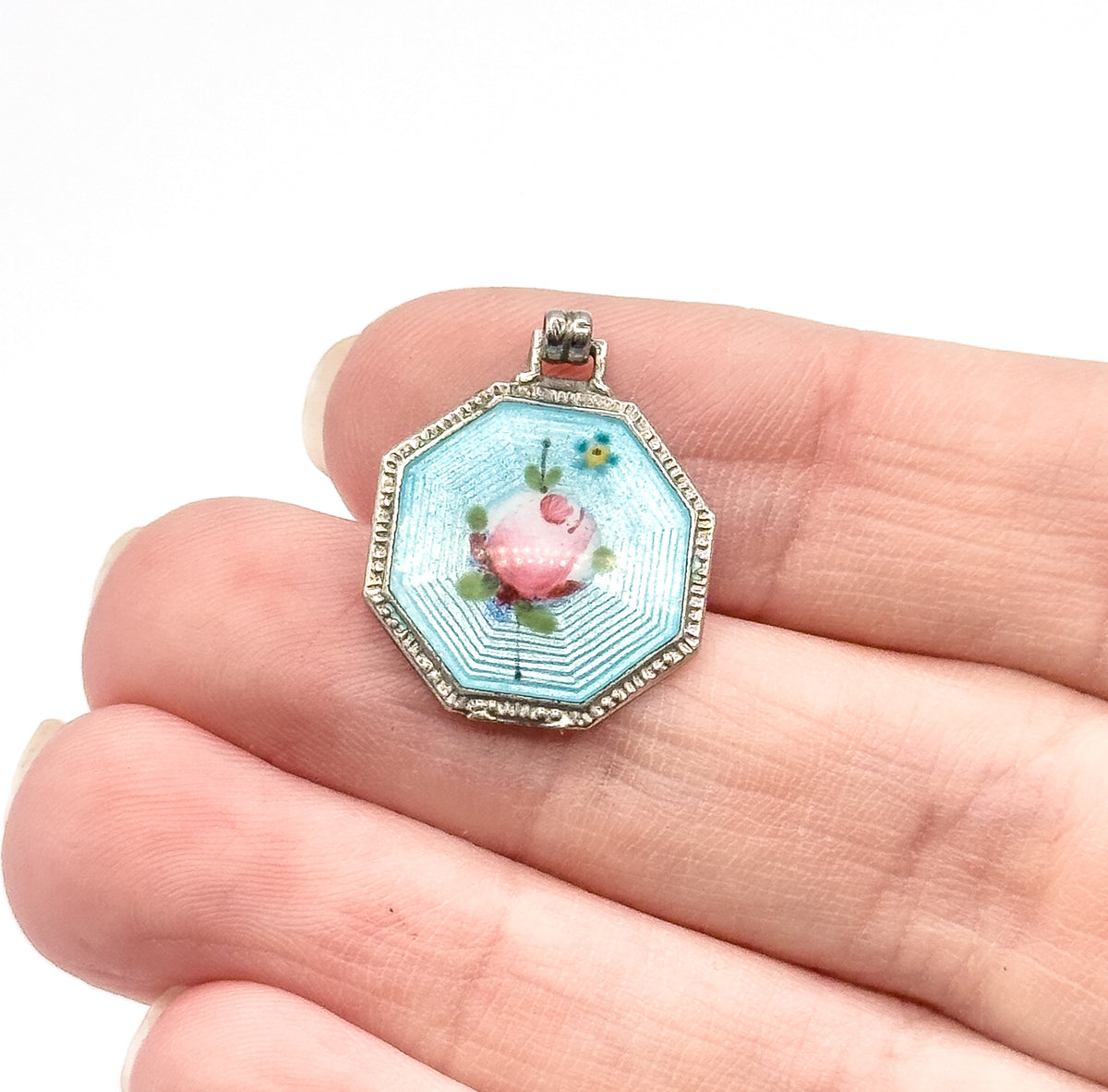Octagonal sterling silver guilloché enamel charm with a pink painted rose on a pale blue field, resting on fingertips that show its small size, its ribbed bail at the top.