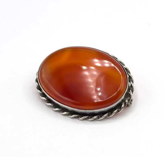 A raised three-quarter view against white, the domed oval carnelian agate catching highlights that reveal its banding, ringed by the twisted sterling rope bezel with darker patina between the strands.