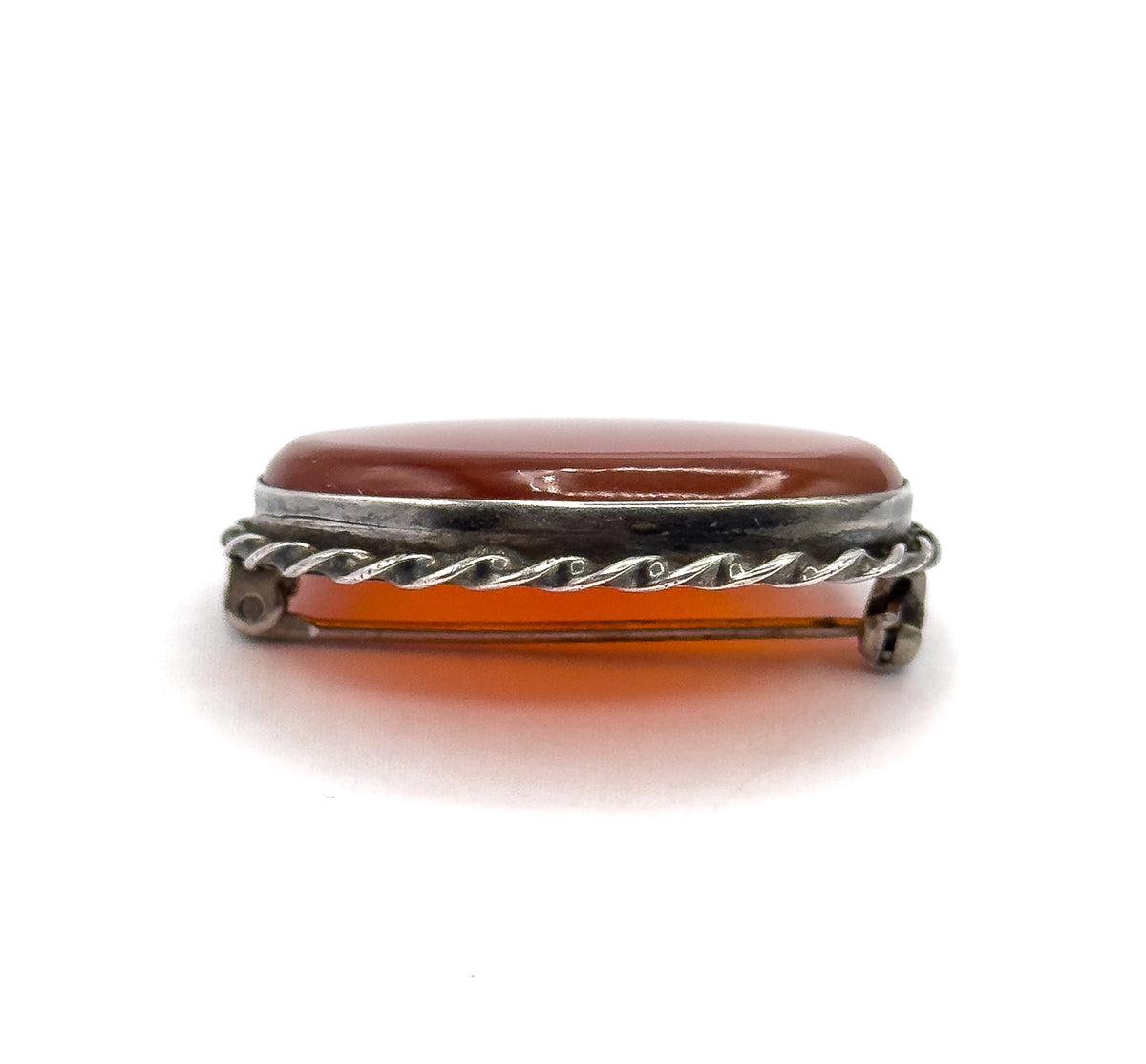A straight side-on edge view against white, the carnelian agate's domed profile rising above the plain bezel rim with the twisted rope below, and the pin stem and catch in silhouette behind.