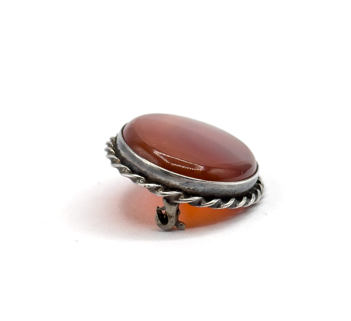 A low tilted view from the front edge, the smooth bezel wall and twisted rope below catching light while the hooked catch juts out at the lower rim and the carnelian agate glows warmly behind it.