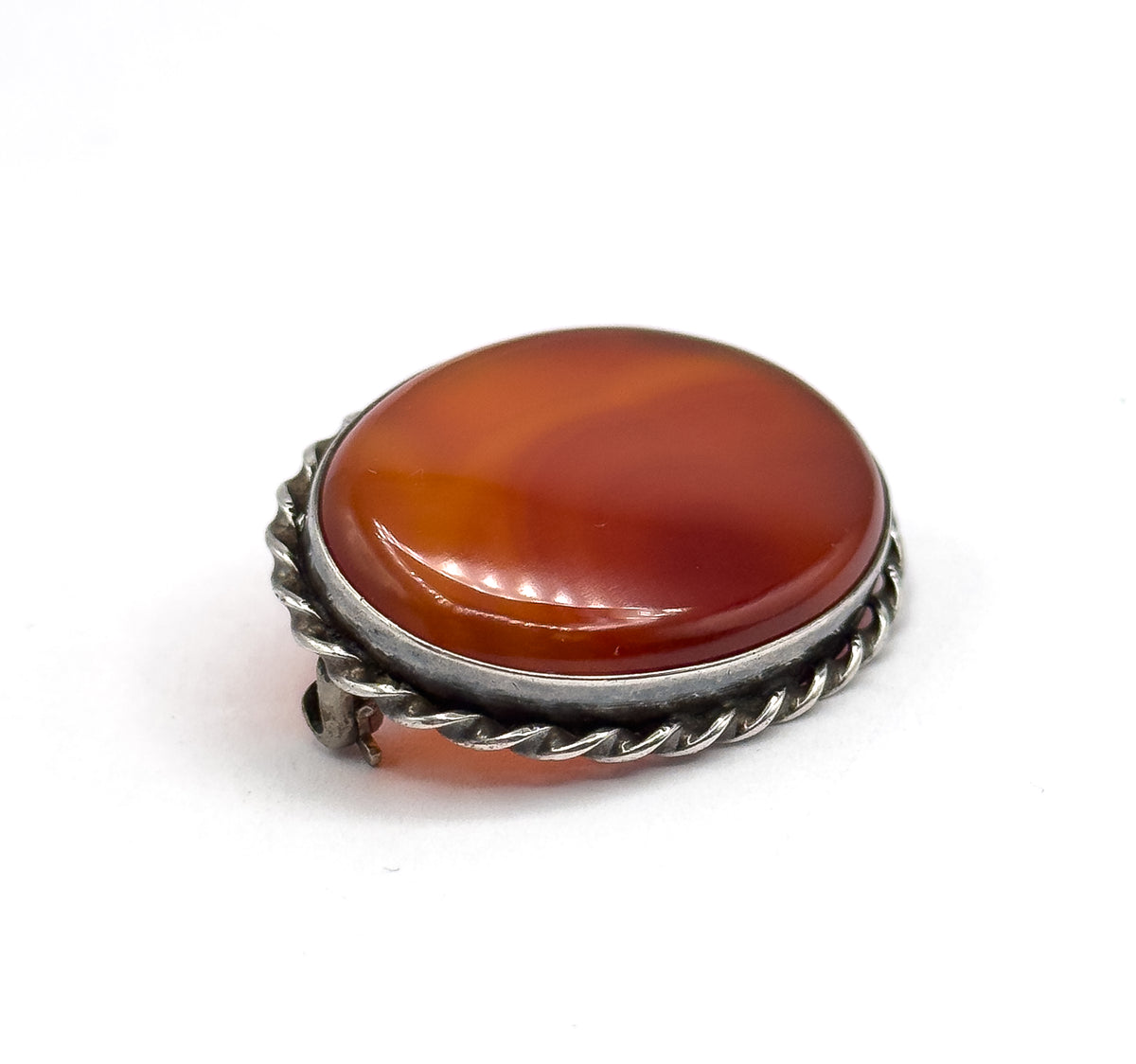 Looking down onto the full oval face at a slight tilt, the banded carnelian agate dominating within its smooth bezel and twisted rope edge, with the pin stem's tip just visible past the left rim.