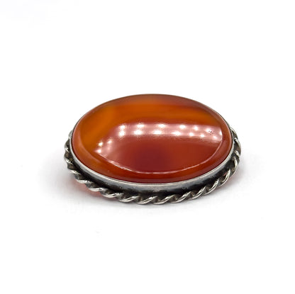 Tilted three-quarter view from above the front edge with no pin fittings in sight, the domed carnelian agate's curved banding and reflections filling the bezel above the twisted rope frame.