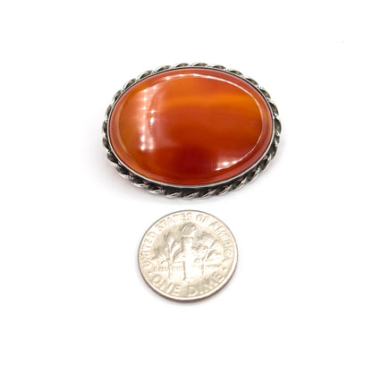The brooch lies face-up beside a dime on white for scale, the banded carnelian agate in its twisted rope frame reading noticeably wider and taller than the coin.