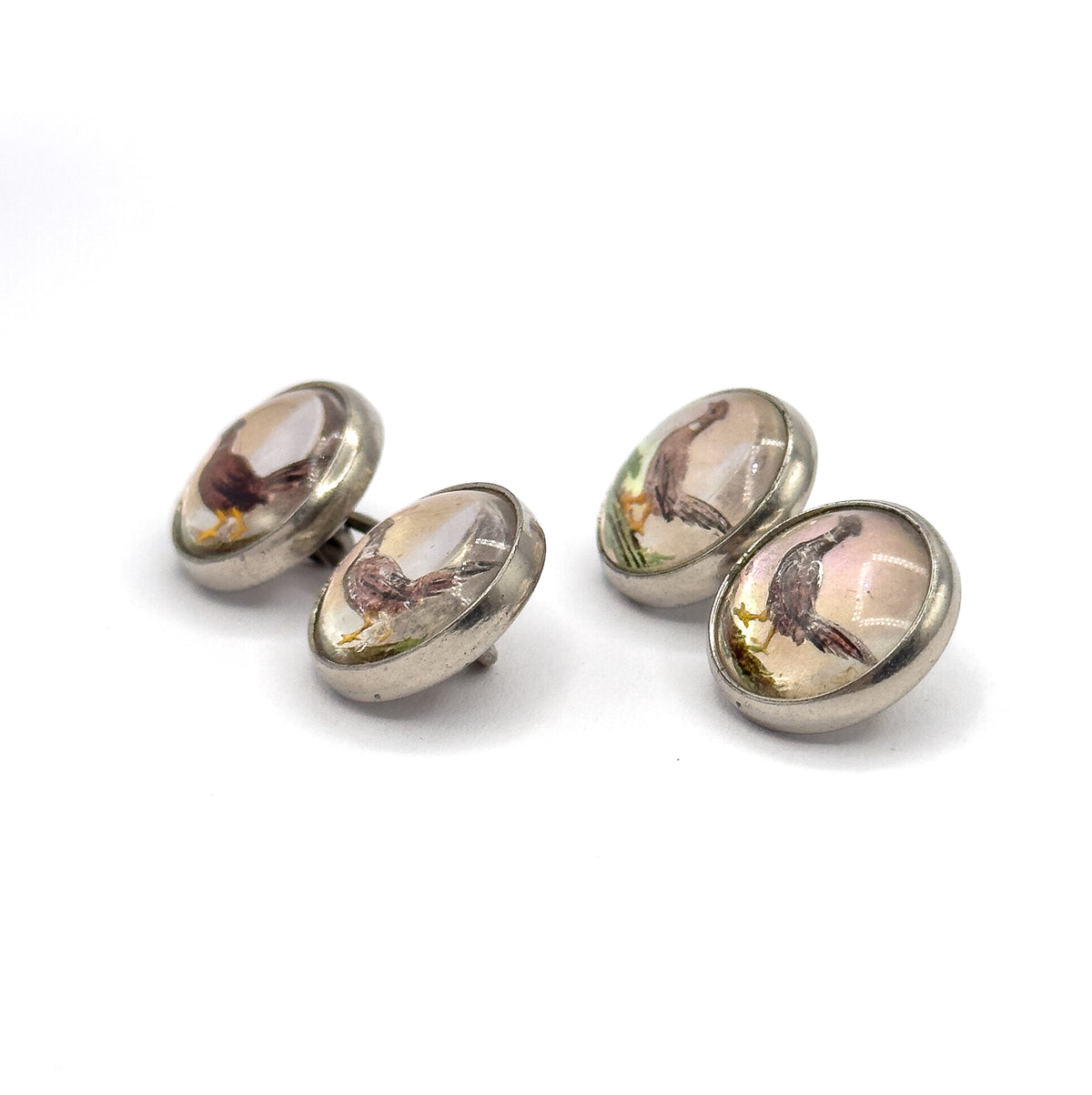 Both cufflinks stand upright on white, all four painted pheasant faces angled towards the camera within their smooth sterling bezels, the domed glass catching soft highlights.