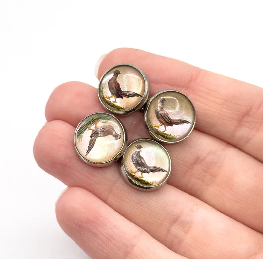 Art Deco reverse painted glass pheasant cufflinks in sterling silver, shown resting on fingers so their four small round faces appear little wider than a fingertip, each bird painted in warm browns.