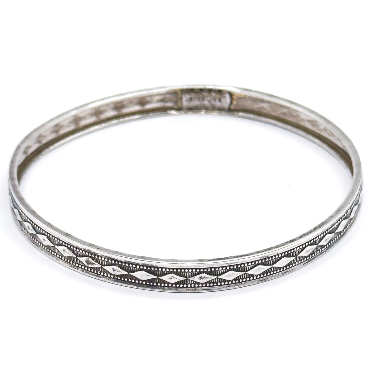 The whole bangle seen from above at a tilt, its full circle in view, with a small stamped mark on the plain inner wall at the far side and the diamond pattern running along the near edge.