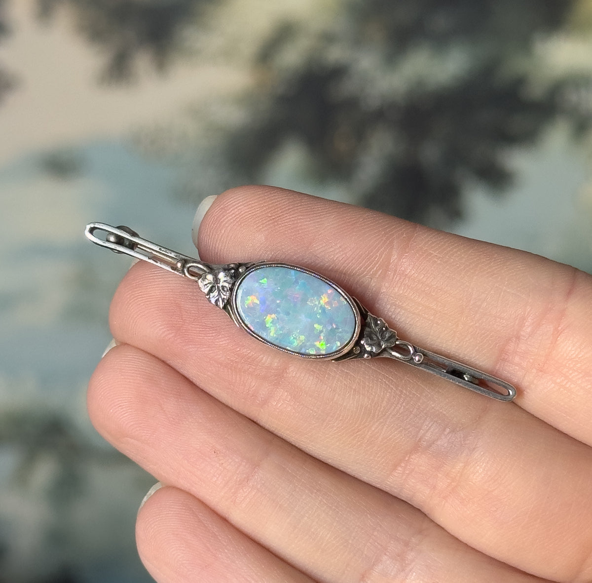 Vintage opal doublet floral brooch in sterling silver, shown resting across fingers for scale, its oval stone flashing blue and green between floral silver mounts on a slender bar with pin stem.