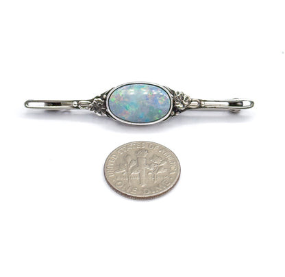 The brooch lies flat beside a dime for scale, its oval opal doublet in a bezel between floral silver mounts spanning a bar about three coin-widths long.